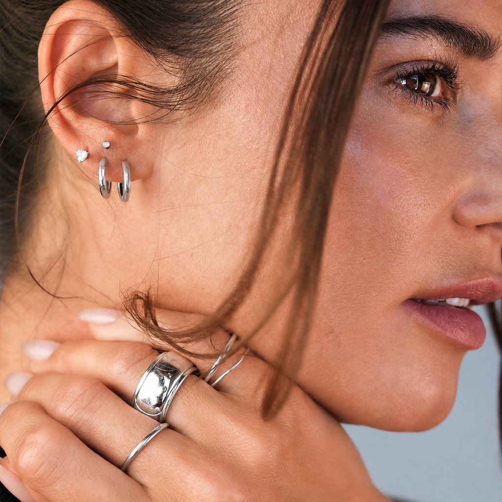 Close-up of a woman wearing multiple silver earrings and rings, including hoop earrings, a small stud, and a wide band ring, with her hand near her neck.