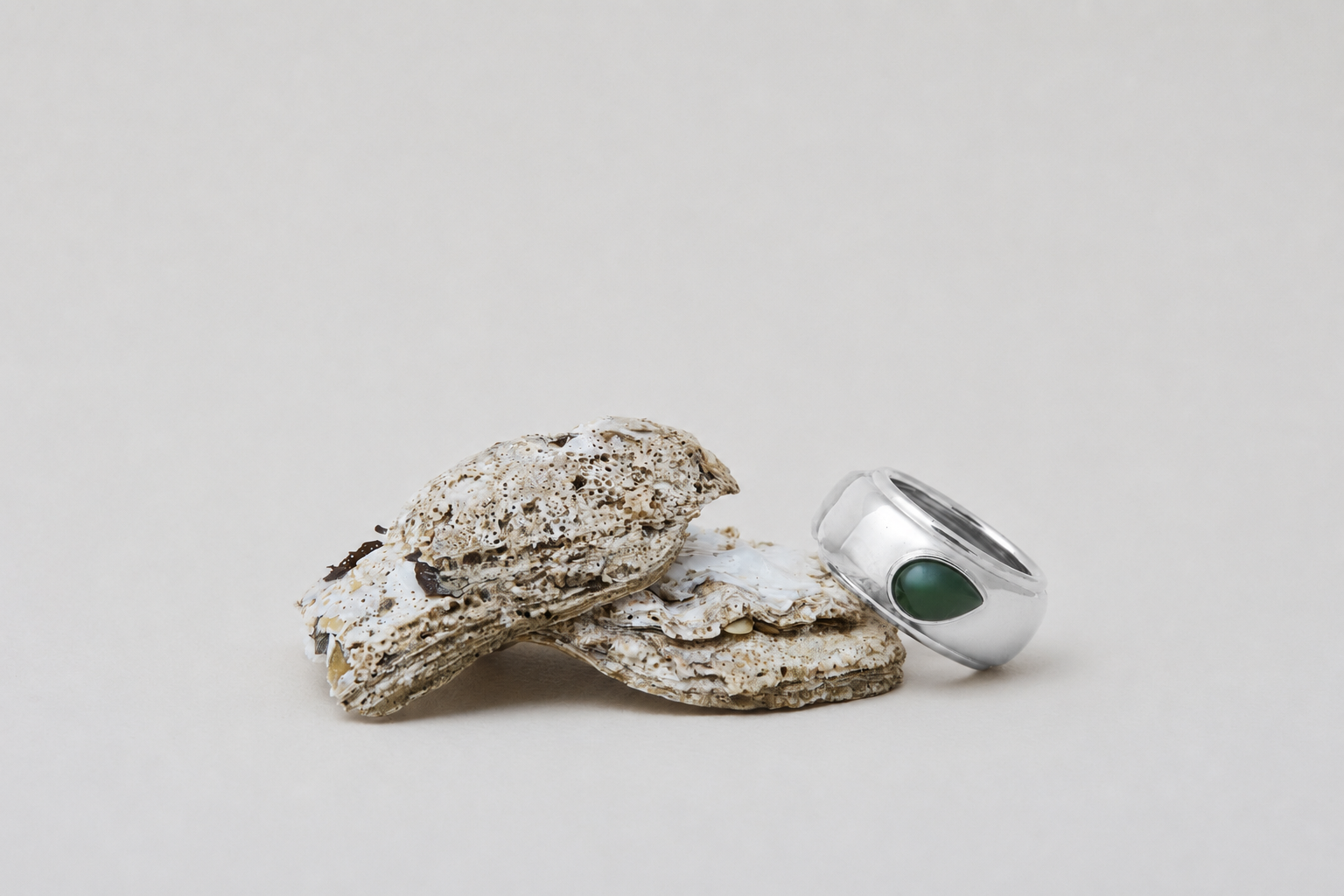 A silver ring with a green teardrop-shaped gemstone, displayed next to three textured, weathered stone pieces on a neutral light gray background.